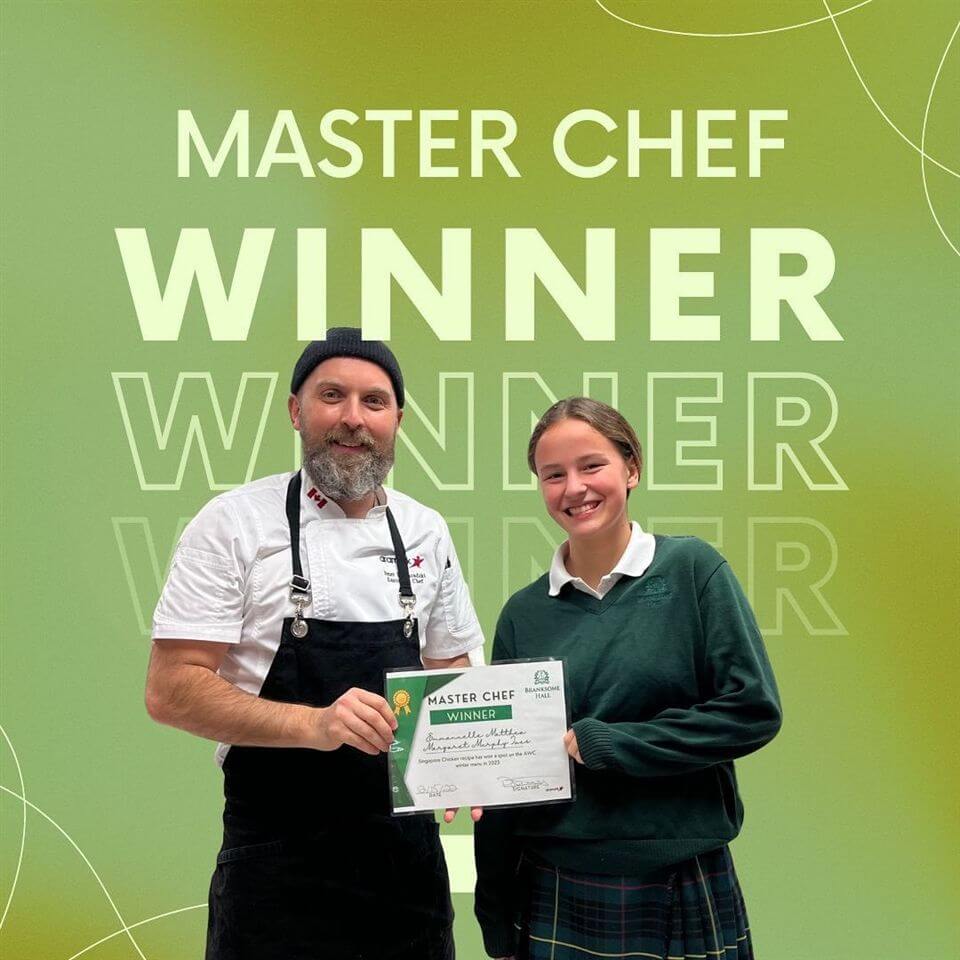 Two people smiling and holding a certificate in front of a green background that says "MASTER CHEF WINNER." One wears a chef's uniform and beanie; the other wears a green sweater and plaid skirt.