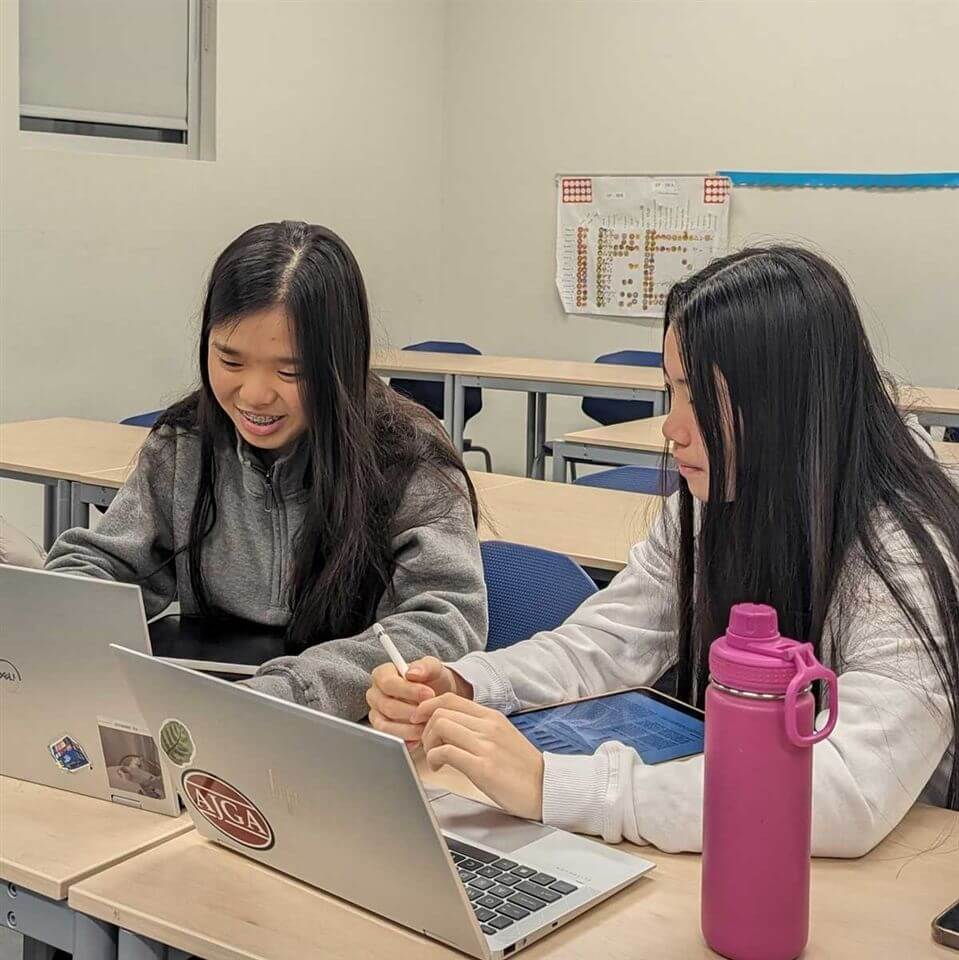 Two girls sit at desks in a classroom, working on laptops and writing on tablets. One is smiling, while the other is focused. A pink water bottle is on the desk, and a chart is visible on the wall behind them.