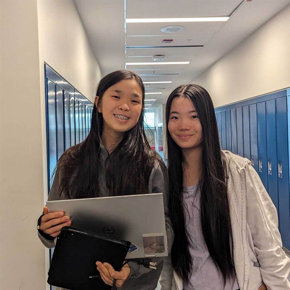 Two young women with long dark hair stand smiling in a school hallway lined with blue lockers. One holds a laptop and a tablet, while the other stands next to her. They both wear casual clothing.