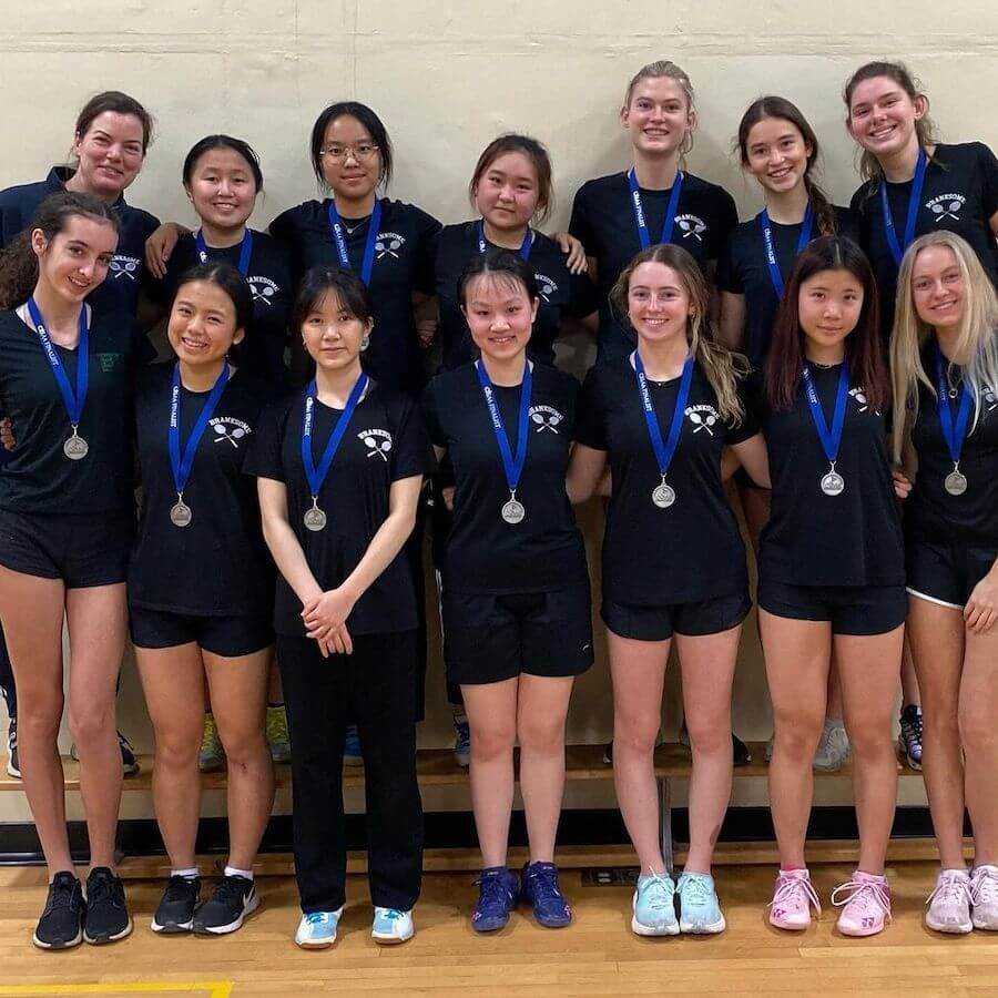 A group of thirteen young women in black sports uniforms stand and kneel indoors, smiling and wearing silver medals around their necks, posing for a team photo against a beige wall.