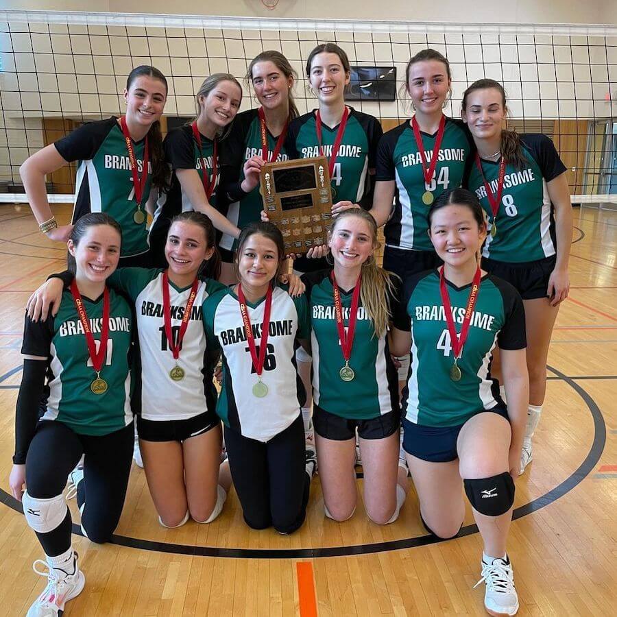 A girls’ volleyball team wearing green and white uniforms poses in a gym with medals around their necks. They are smiling and holding a plaque, celebrating their victory in front of a volleyball net.