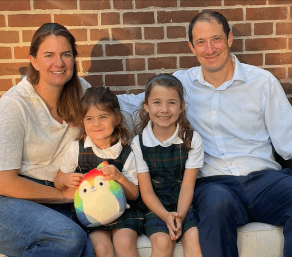 A family of four sits on a couch outdoors, smiling. Two young girls wearing green plaid uniforms sit between their parents. One girl holds a colorful plush toy. A brick wall is in the background.