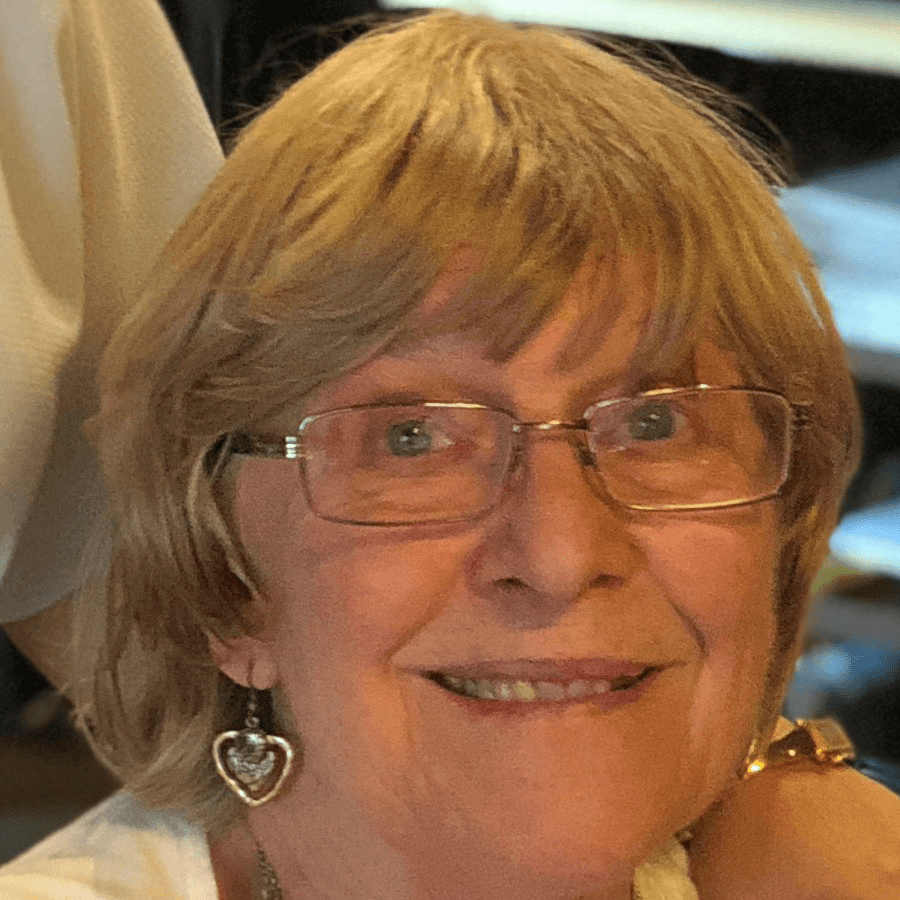 An older woman with short, light brown hair and glasses smiles at the camera. She is wearing heart-shaped earrings and a white top. The background is softly blurred.