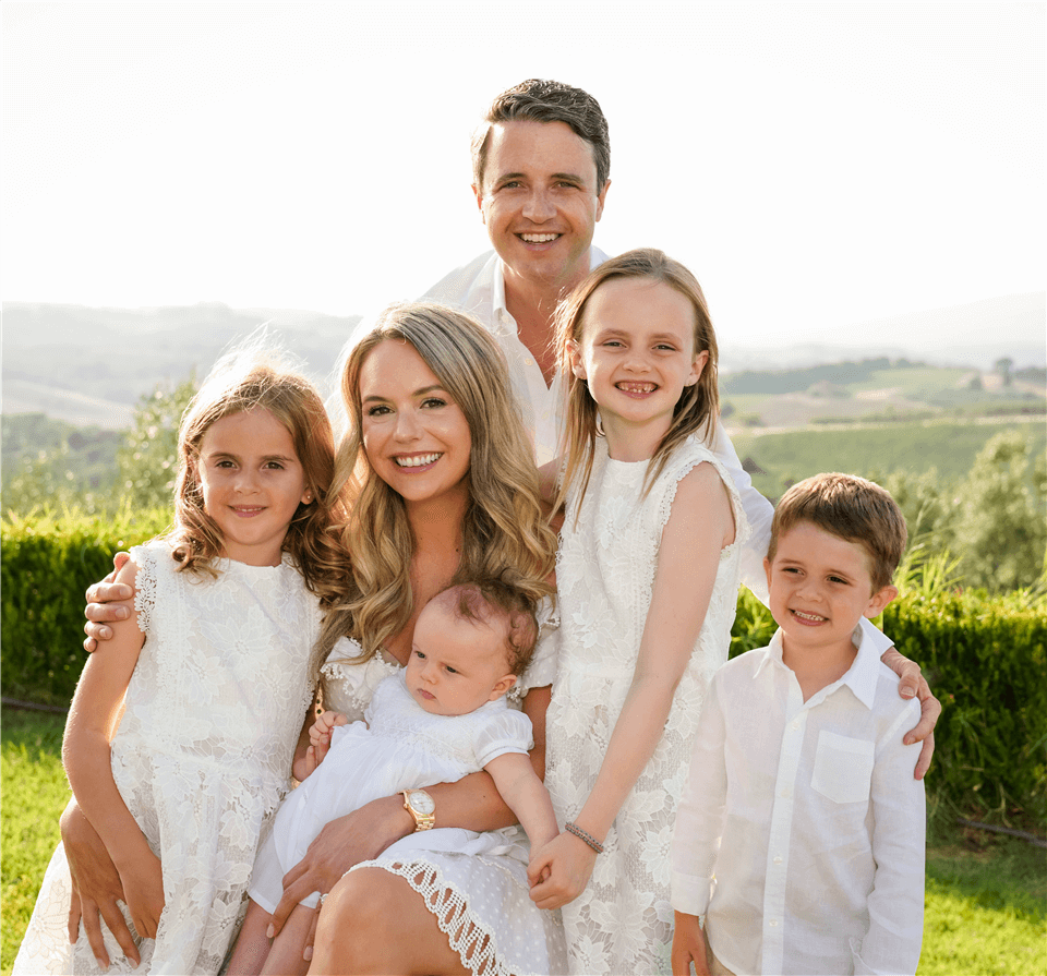A smiling family of five poses outdoors. Two adults and three children stand together, all wearing white clothing, with greenery and hills in the background. The woman holds a baby, and everyone looks happy.