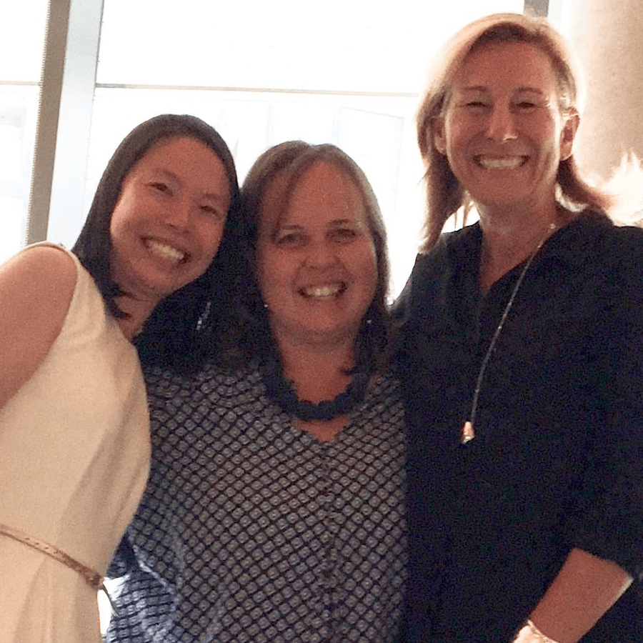 Three women smiling and posing closely together indoors. One woman is wearing a white dress, another a patterned blouse with a blue necklace, and the third a dark top. They appear happy and are standing in front of large windows.