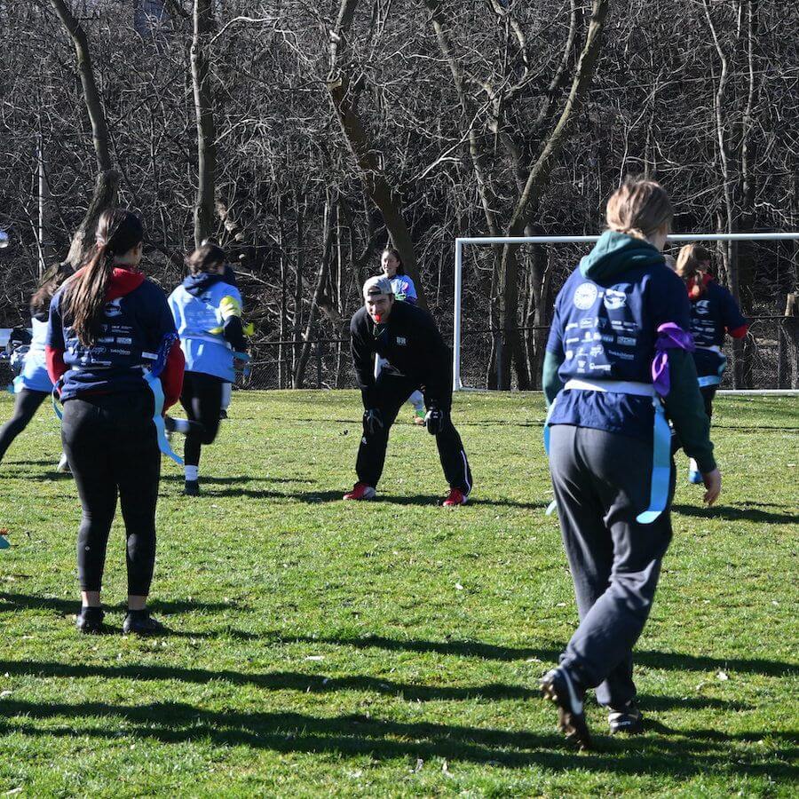 A group of people play soccer on a grassy field, with some wearing blue vests. Leafless trees are visible in the background, and a metal goalpost stands behind the players. The weather appears sunny and cold.