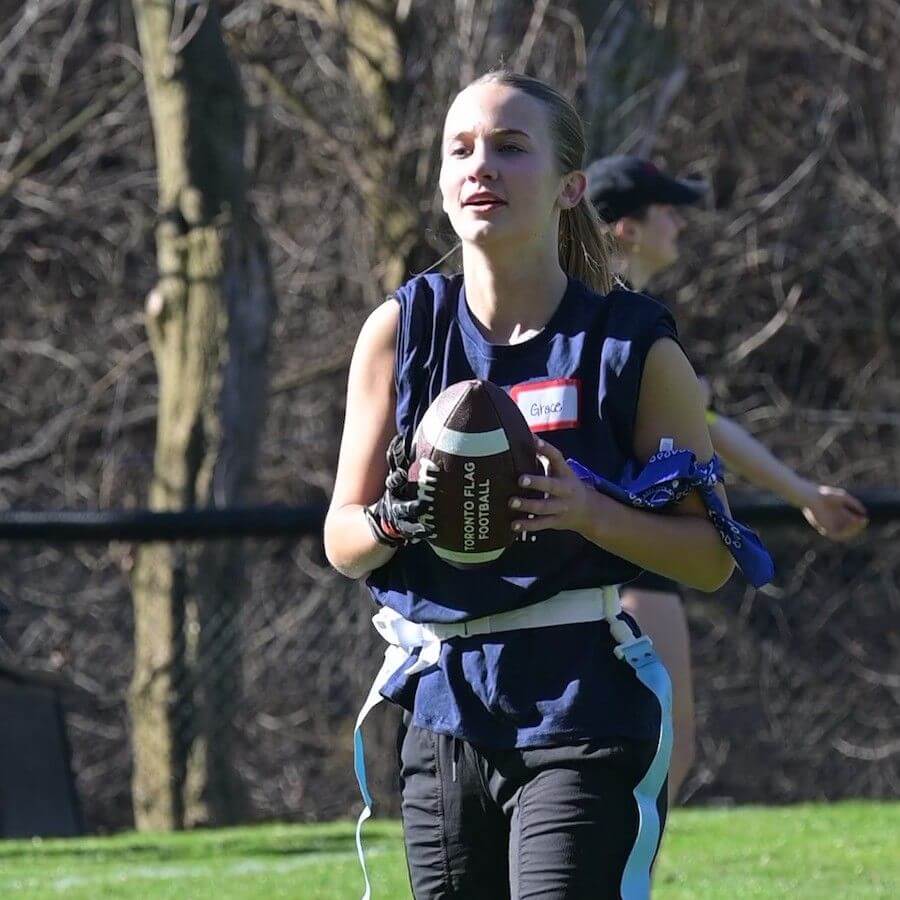 A girl wearing a navy-blue sleeveless shirt, flag football belts, and gloves holds a football while standing on a grassy field with trees in the background. Another player is visible in the distance.