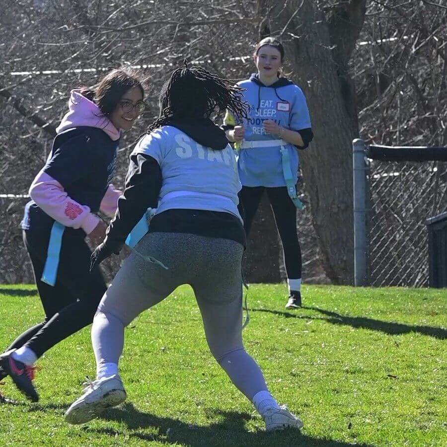 Three people play flag football on a sunny grassy field; one runs with a flag belt as another tries to grab it, while a third person stands watching in the background. Trees and a fence are visible behind them.