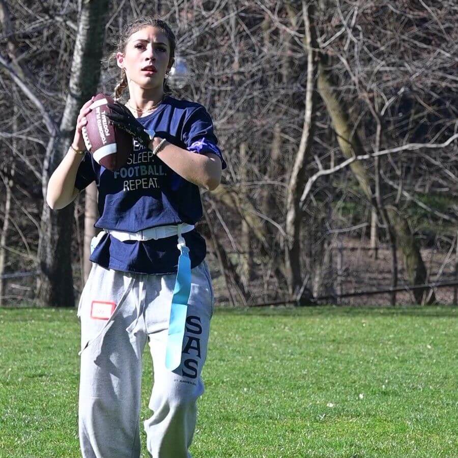 A young woman outdoors holding a football, wearing a navy t-shirt and gray sweatpants with a flag belt, preparing to throw during a flag football game. Trees and grass are visible in the background.