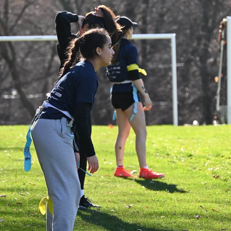 Three people stand on a grassy field, wearing athletic clothing and flag football belts, appearing focused and ready to play, with a goalpost and bare trees in the background.