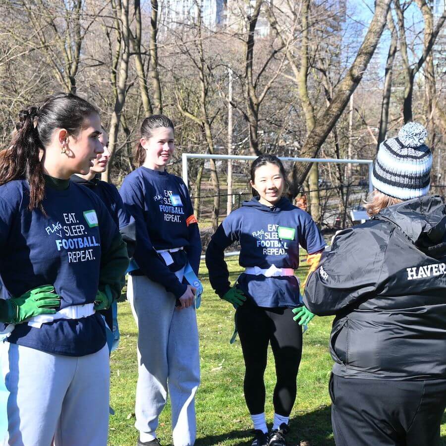Four young women in matching "Eat. Sleep. Football. Repeat." shirts stand in a circle, smiling and listening to a coach in a "HAVERFORD" jacket, on a sunny field with trees and a soccer goal in the background.