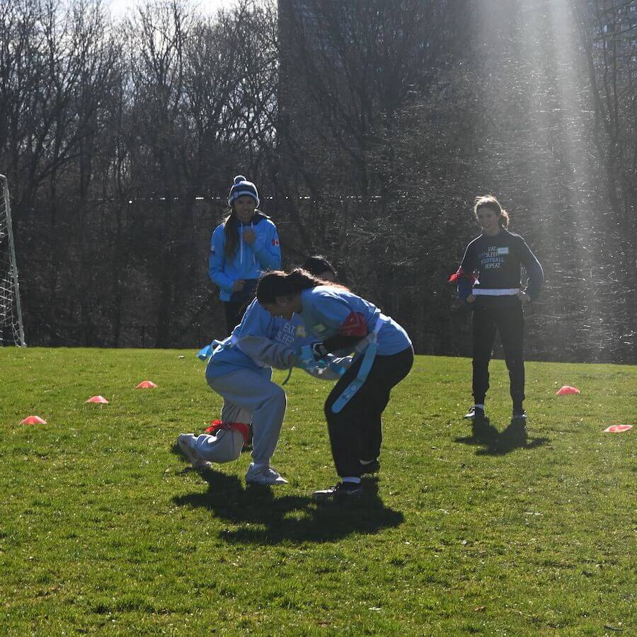 Four people are on a grassy field; two are engaged in a tackle, while two others watch. Orange cones mark the area, and sunlight beams down from the top right. Leafless trees are visible in the background.