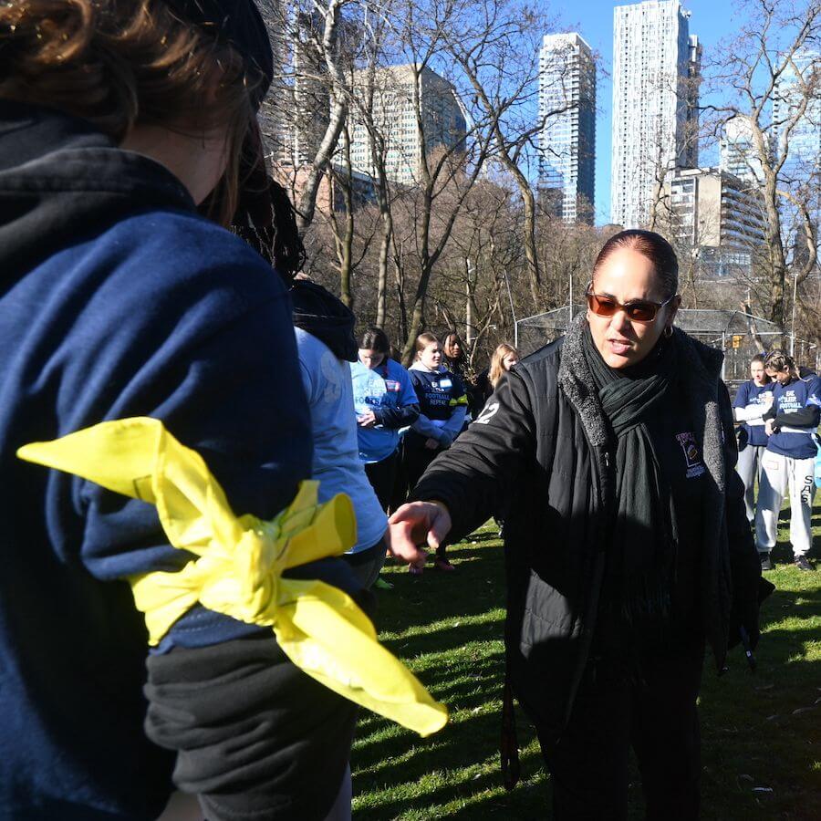 A woman in sunglasses points as she talks to a person with a yellow band tied around their arm. They are outdoors in a park with other people and city buildings in the background.
