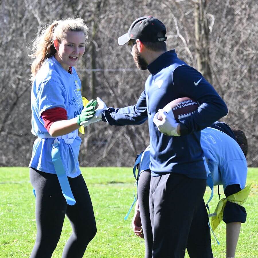 Two people wearing blue shirts and flag football belts interact on a grassy field; one holds a football while the other smiles and collects a flag. Another person in the background is bent over, adjusting their belt.