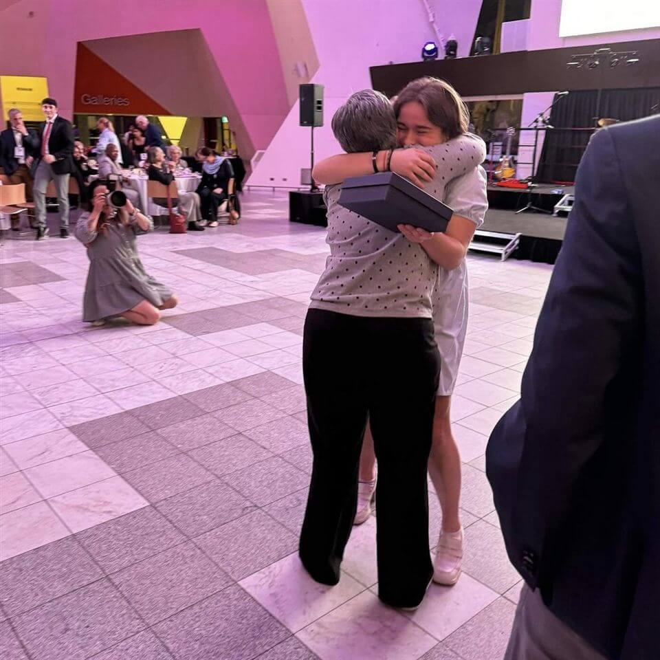 A young person in a white outfit hugs an older woman while holding a box, surrounded by people in a large indoor space. A photographer kneels to capture the moment, and others watch in the background.