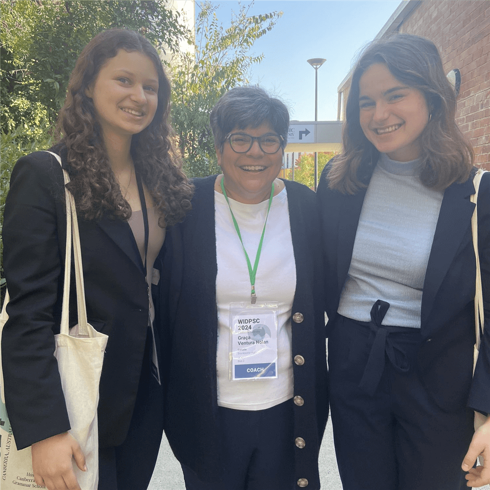 Three women stand together outdoors, smiling for the camera. Two younger women wear dark blazers, while the woman in the center wears glasses, a white shirt, and a conference badge with a green lanyard. Trees and buildings are visible.