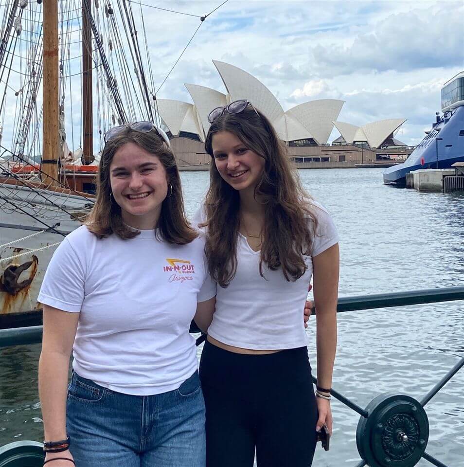 Two young women smiling and posing by the waterfront, with the Sydney Opera House and docked boats visible in the background under a partly cloudy sky.