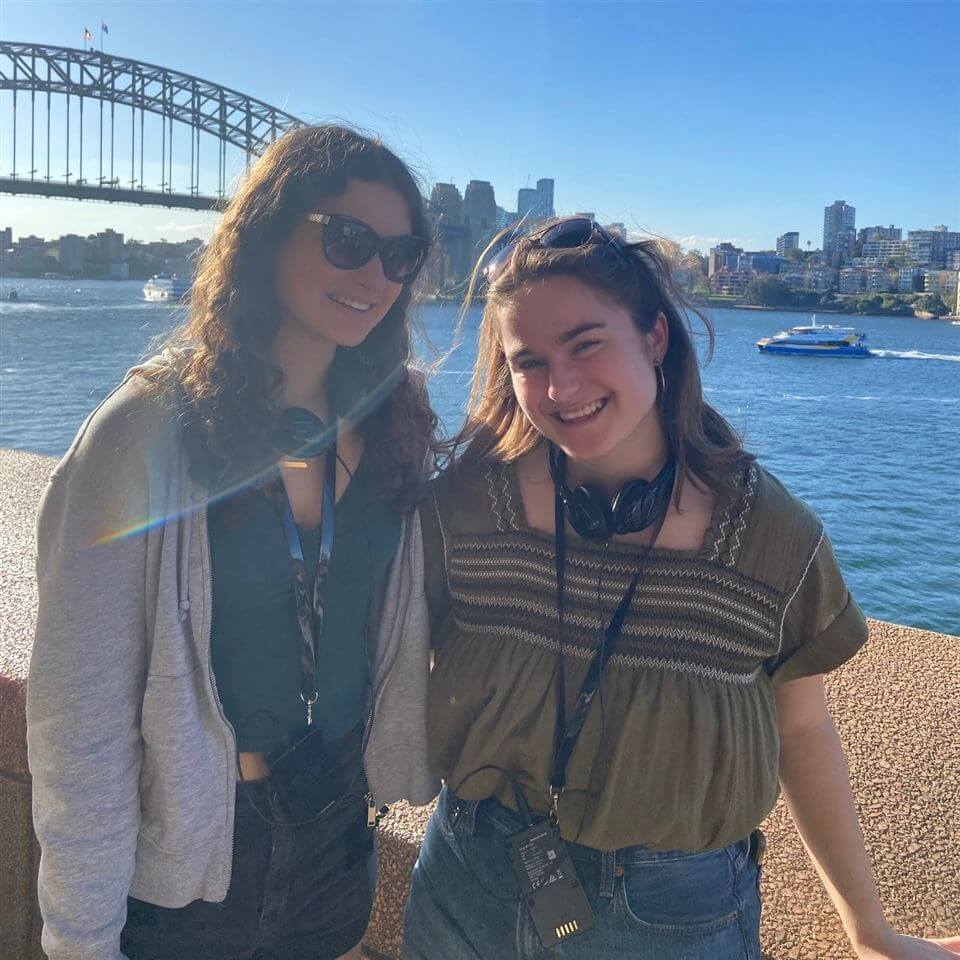 Two young women smiling and posing together outdoors by the water, with the Sydney Harbour Bridge and city skyline in the background on a sunny day.