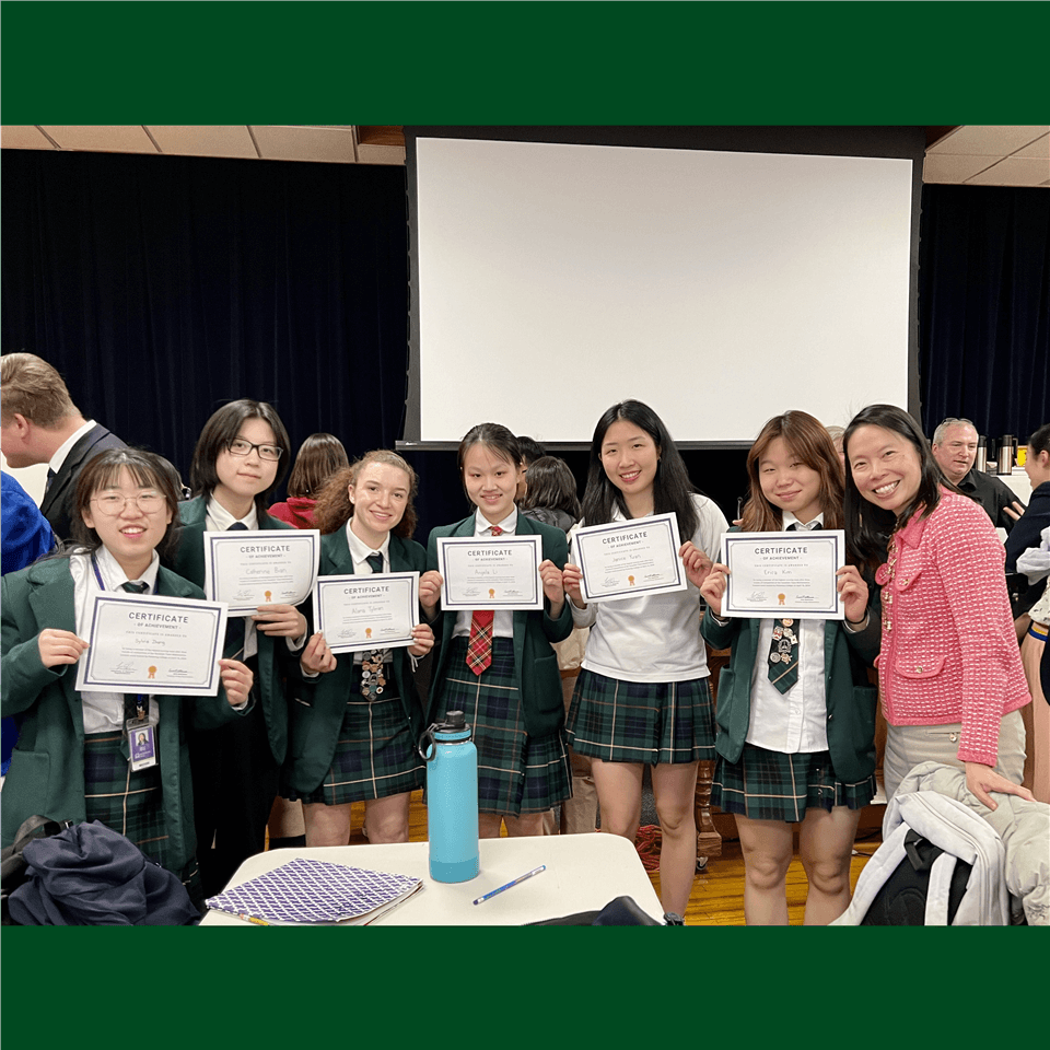A group of students in school uniforms stand together indoors, smiling and holding certificates. An adult stands at the end of the group, also smiling. Other people and tables are visible in the background.
