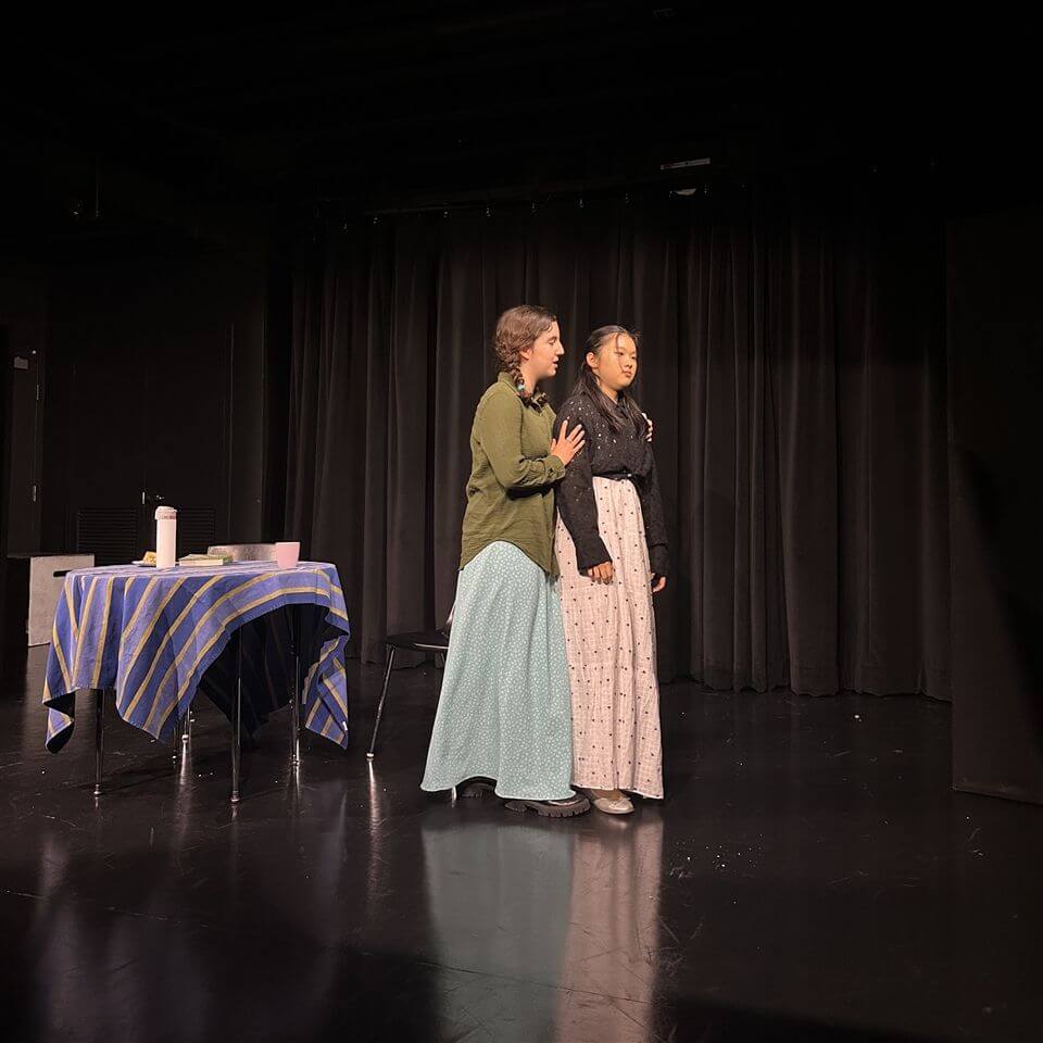 Two young women in long skirts stand close together on a dark stage, one comforting the other. A table covered with a blue-striped cloth is on the left, and black curtains hang in the background.