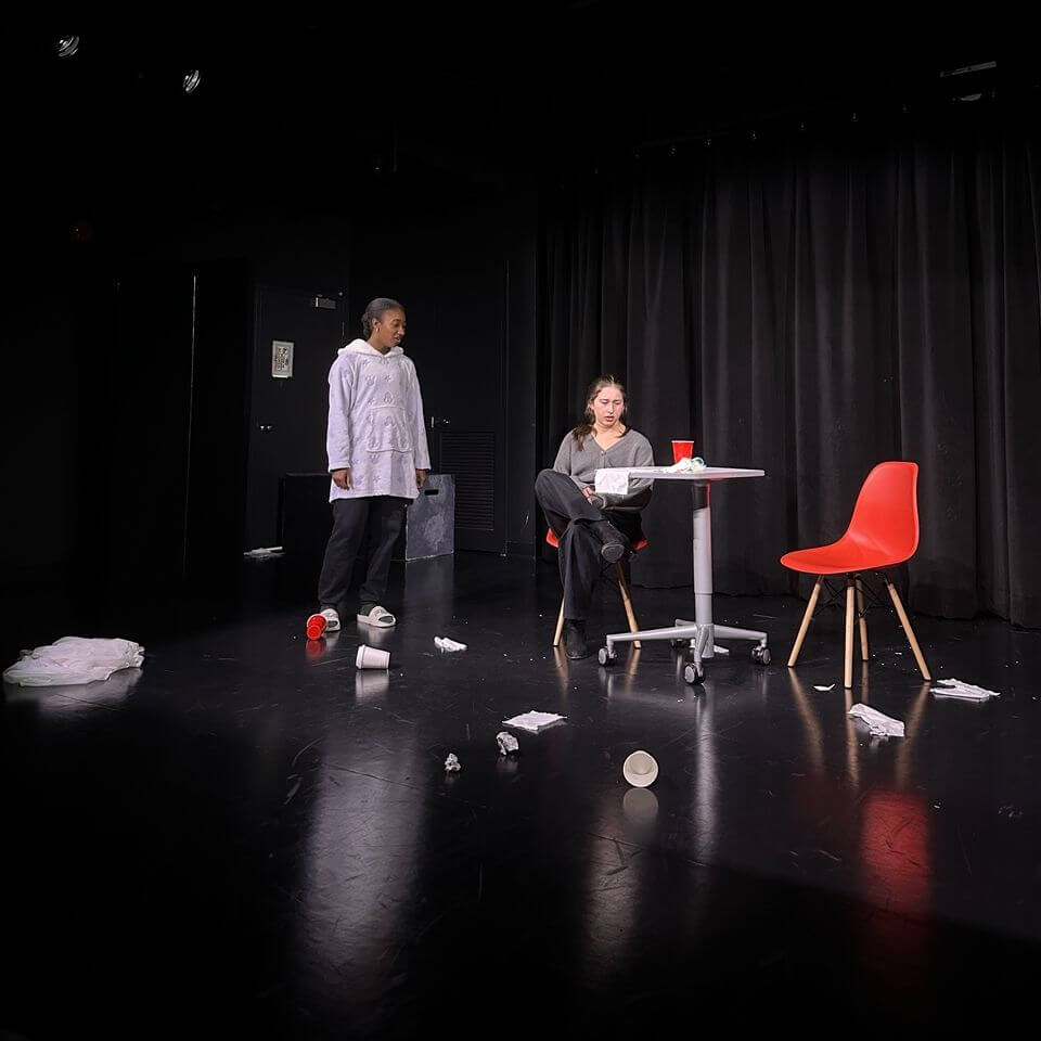 Two people perform on a dark stage; one stands wearing a white coat, the other sits at a table with red chairs. Crumpled papers and cups are scattered on the floor, with black curtains in the background.