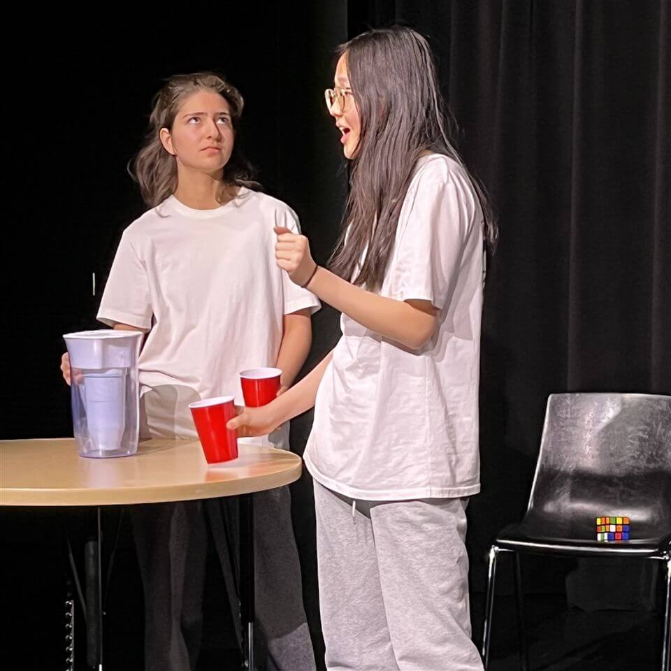 Two young women in white t-shirts and gray pants stand by a table with a water pitcher and red cups. One speaks animatedly while the other looks at her with a skeptical expression. Black chairs and a dark backdrop are behind them.
