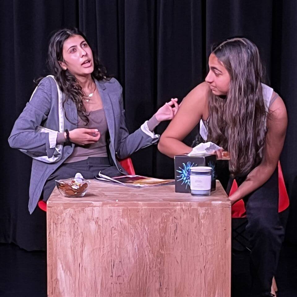 Two women sit at a wooden table on stage. One gestures animatedly while talking, holding a white bag, as the other looks down, reaching for a tissue box. Both appear engaged in a dramatic conversation. Black curtain backdrop.