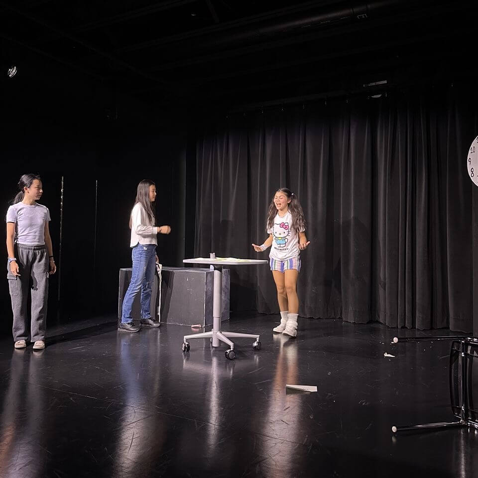Three young women perform on a dark stage with black curtains, standing around a small round table on wheels. One woman wears colorful shorts and white boots, while the others wear casual pants and tops. Props are scattered on the floor.