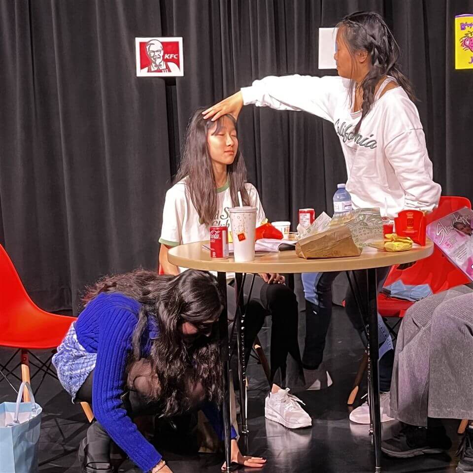 Three girls are on stage. One sits at a table with fast food, eyes closed, while another stands and touches her head. A third girl kneels on the floor in front of them. Black curtains and KFC sign are in the background.