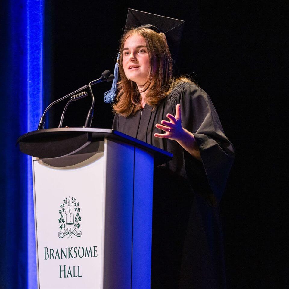 A student in a graduation cap and gown speaks at a podium labeled "Branksome Hall," gesturing with one hand, under stage lighting with a blue curtain in the background.
