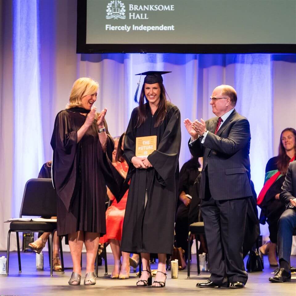 A female graduate in a cap and gown smiles on stage holding a book labeled "The Future" while a woman and a man on either side applaud her during a graduation ceremony. Branksome Hall branding appears on a screen above.