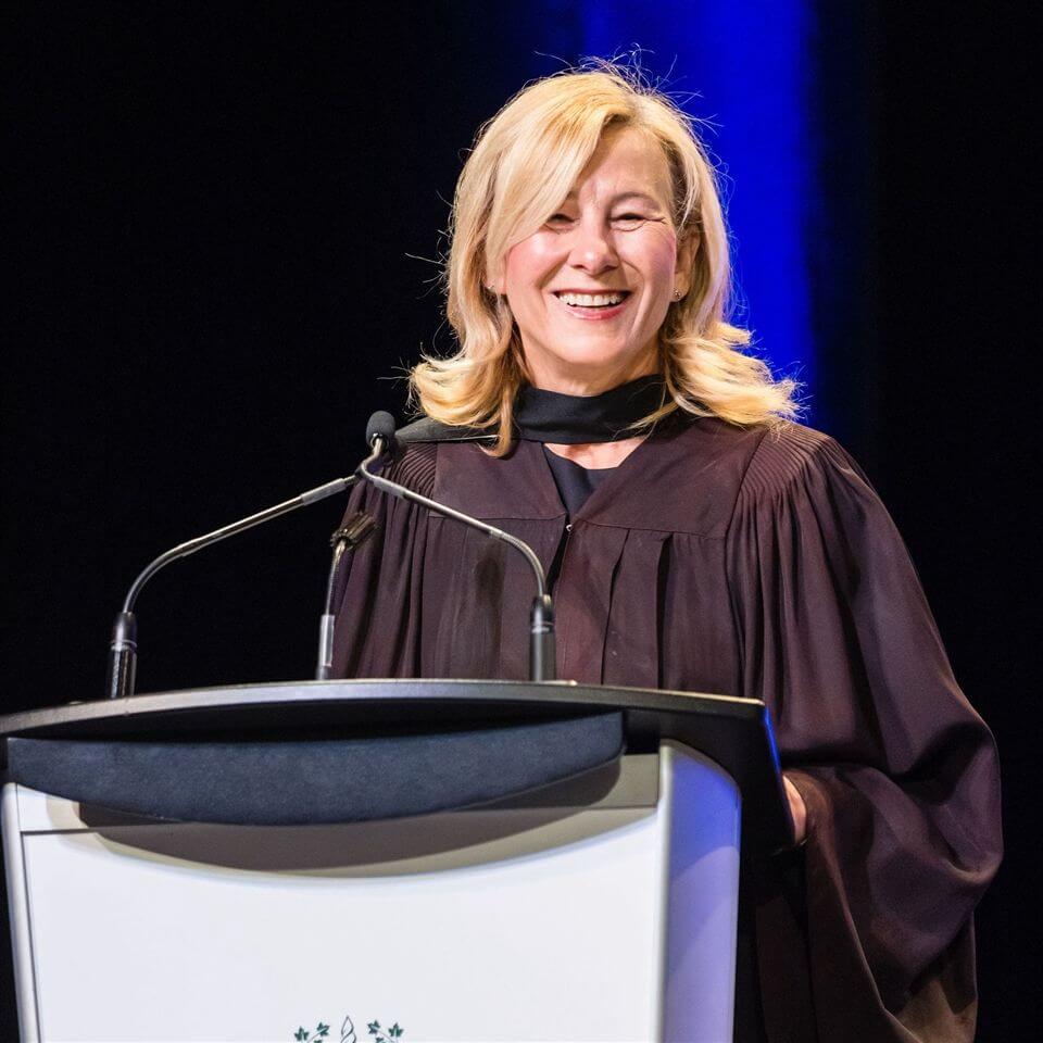 A woman in academic robes smiles while speaking at a podium with microphones during a graduation ceremony. The background is dark with a blue vertical light.