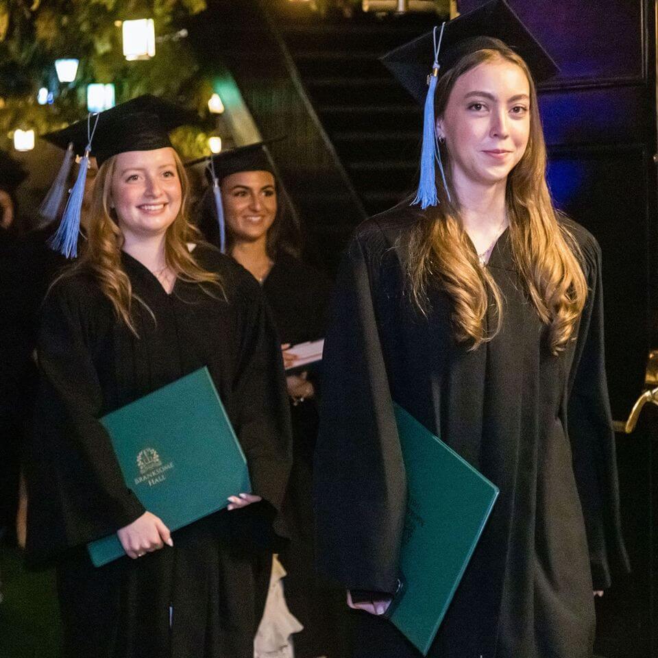 Three young women in black graduation gowns and caps hold green diploma folders, smiling as they walk indoors during a graduation ceremony. Warm lighting and blurred decorations are visible in the background.