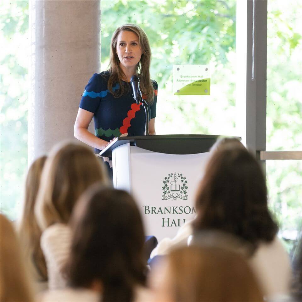 A woman stands at a podium with a microphone, speaking to an audience. The podium displays the Branksome Hall logo. Sunlight streams through large windows, revealing green foliage outside.