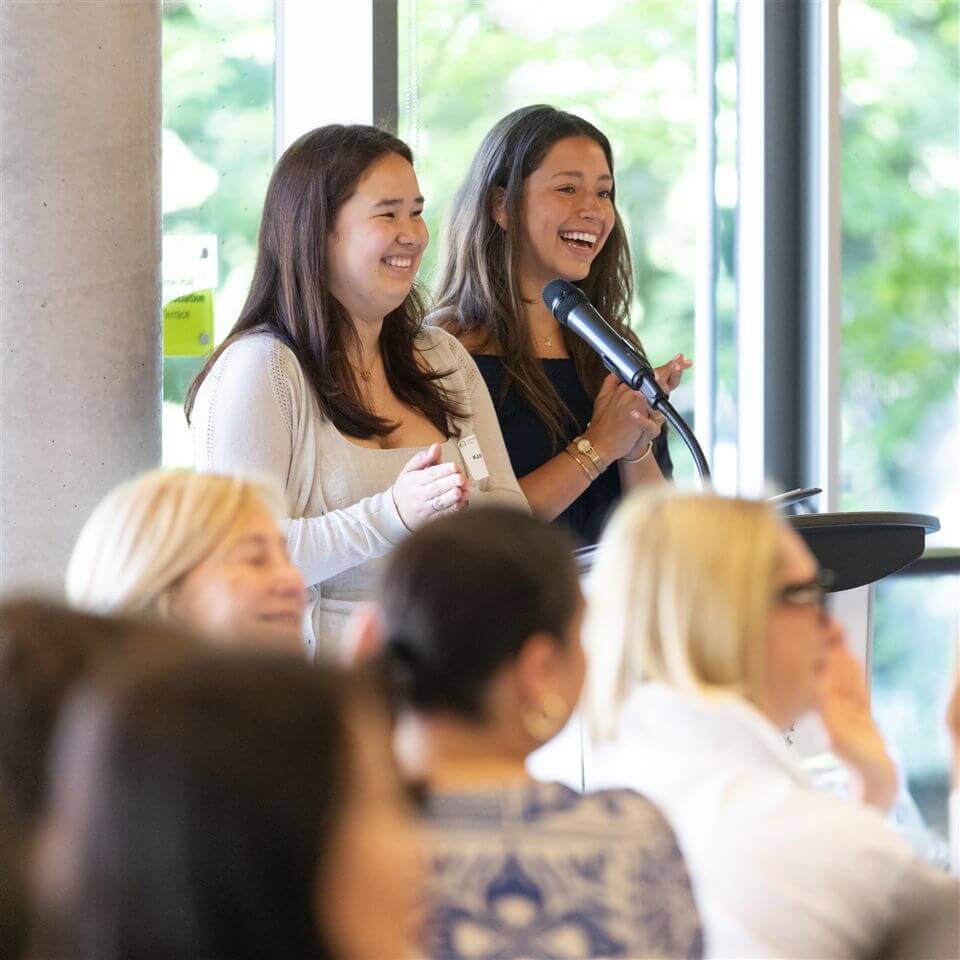 Two women stand at a podium, smiling and clapping, while speaking to an audience in a bright room with large windows and greenery visible outside.