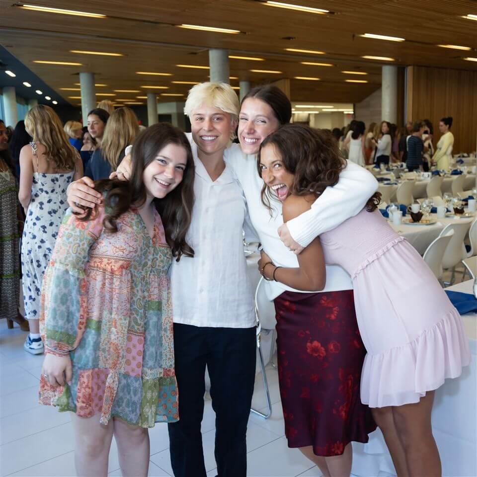 Four people stand close together and smile at the camera at an indoor event with tables set for a meal behind them. The group includes three women in colorful dresses and one person in a white shirt and black pants.