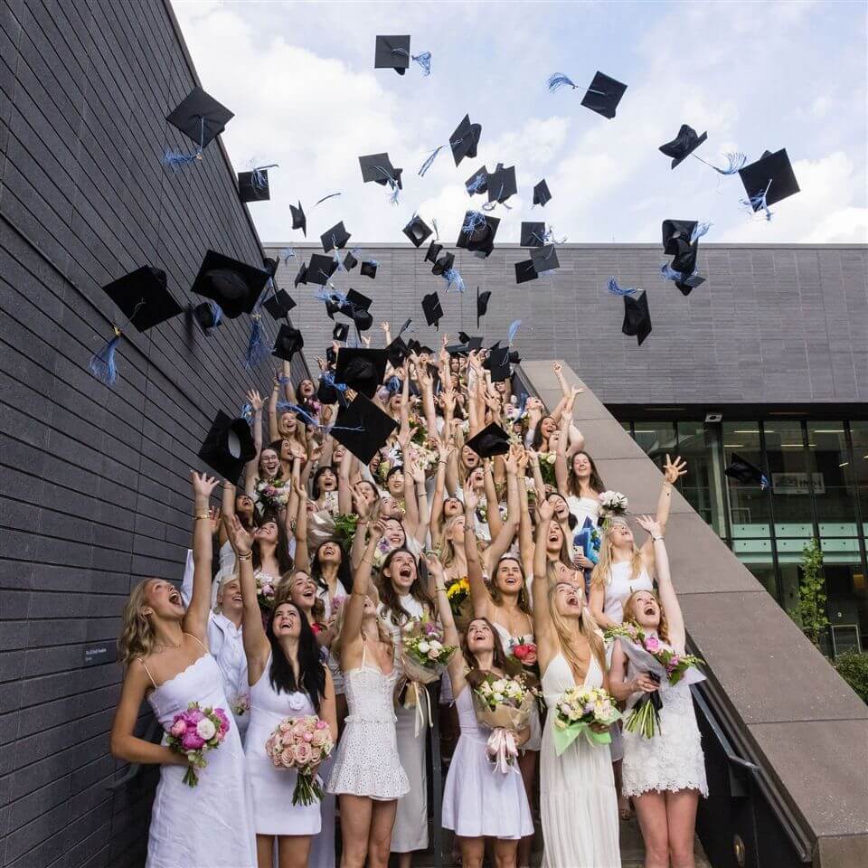 A large group of graduates in white dresses toss their caps into the air on an outdoor staircase, celebrating with flowers and joyful expressions against a modern building backdrop.