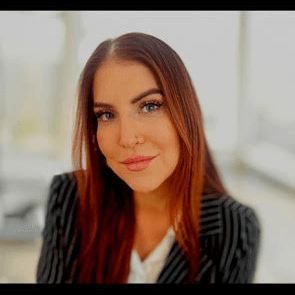 A woman with long reddish-brown hair, wearing a black pinstripe blazer and white blouse, smiles at the camera. She has a nose ring and is in a bright, modern indoor setting.