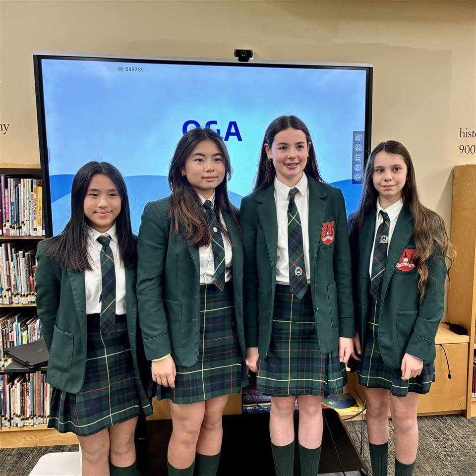 Four girls in green plaid school uniforms stand in front of a large screen displaying "Q&A" in a library setting with bookshelves in the background.