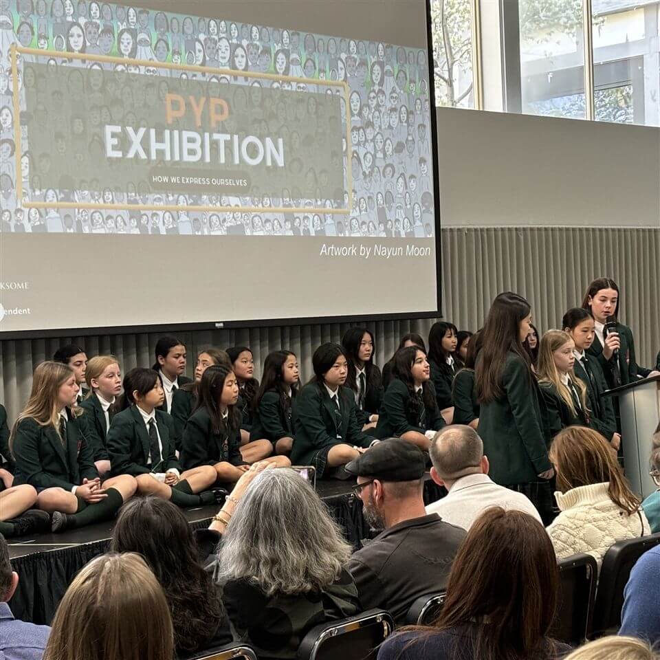 A group of students in green blazers sit and stand on a stage, presenting at a "PYP Exhibition" event to an audience. A large screen behind them displays the event name and the phrase "How We Express Ourselves.