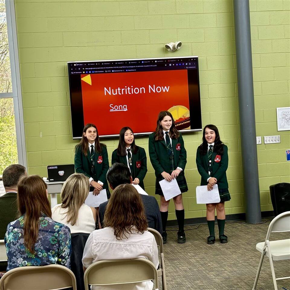 Four girls in green school uniforms stand in front of an audience, presenting in a classroom. Behind them, a screen displays the title “Nutrition Now” and a link labeled “Song.” The audience is seated and listening attentively.