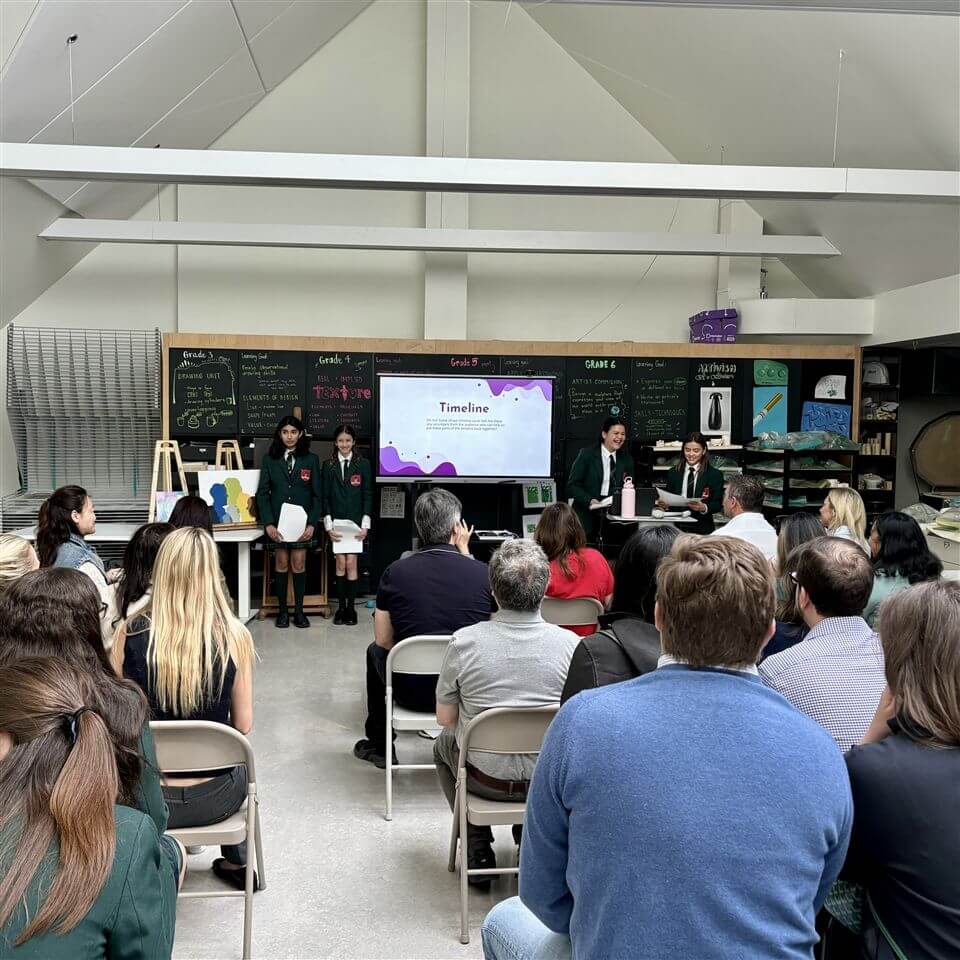 Students in uniforms present a slideshow titled "Timeline" to an audience seated in a classroom with a chalkboard, artwork, and science displays in the background.