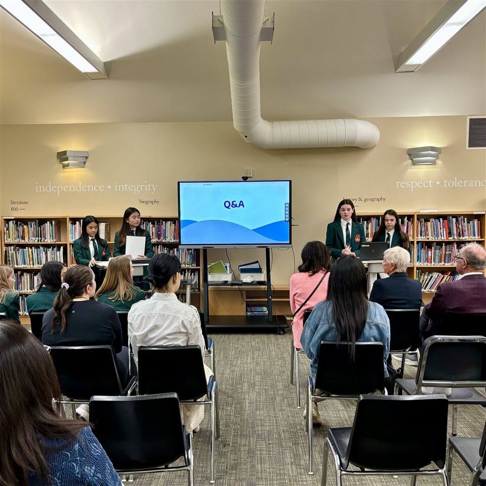 A group of students in green blazers stand at the front of a library, presenting to an audience. A screen displays "Q&A." Bookshelves and words like "independence" and "integrity" are visible on the walls.