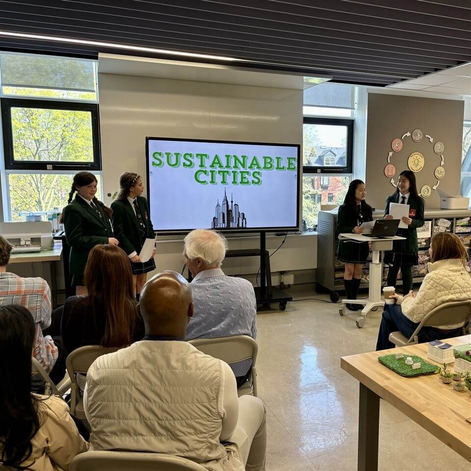 Four students in uniforms present a project titled “Sustainable Cities” on a large screen to an audience seated in a classroom or conference room with large windows and educational displays.