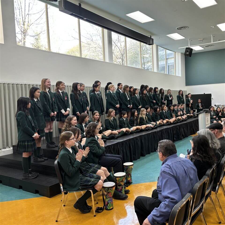 A group of students in green blazers and plaid skirts or pants stand and sit on risers in a school auditorium. Some students in front play drums. Adults sit in the audience, watching the performance.