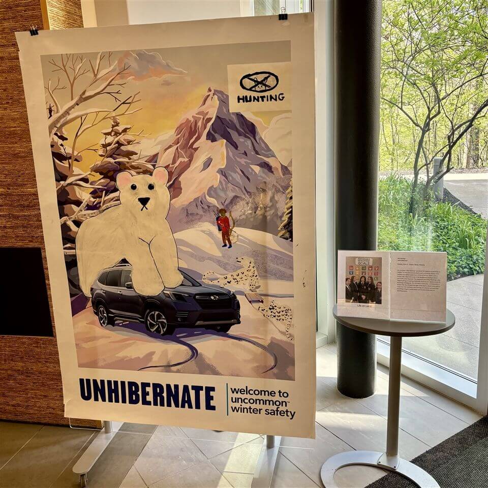 A poster shows a large illustrated polar bear in front of a Subaru car on a snowy mountain road, with a person in red gear and winter trees. Text reads “UNHIBERNATE—welcome to uncommon winter safety.”.