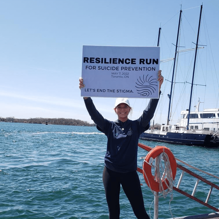 A smiling person stands on a boat, holding up a sign that reads: "Resilience Run for Suicide Prevention, May 7, 2022, Toronto, ON. Let’s end the stigma." Water and boats are visible in the background.