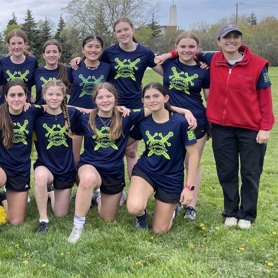 A girls’ sports team poses outdoors on grass, wearing matching navy shirts with neon yellow logos. One adult in a red jacket stands to the side. Trees and buildings are visible in the background.