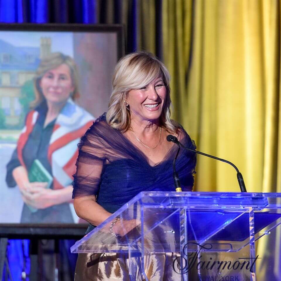 A smiling woman in an elegant outfit stands at a clear podium with microphones, speaking at an event. Behind her is a painted portrait of herself. The background features blue and yellow drapes.