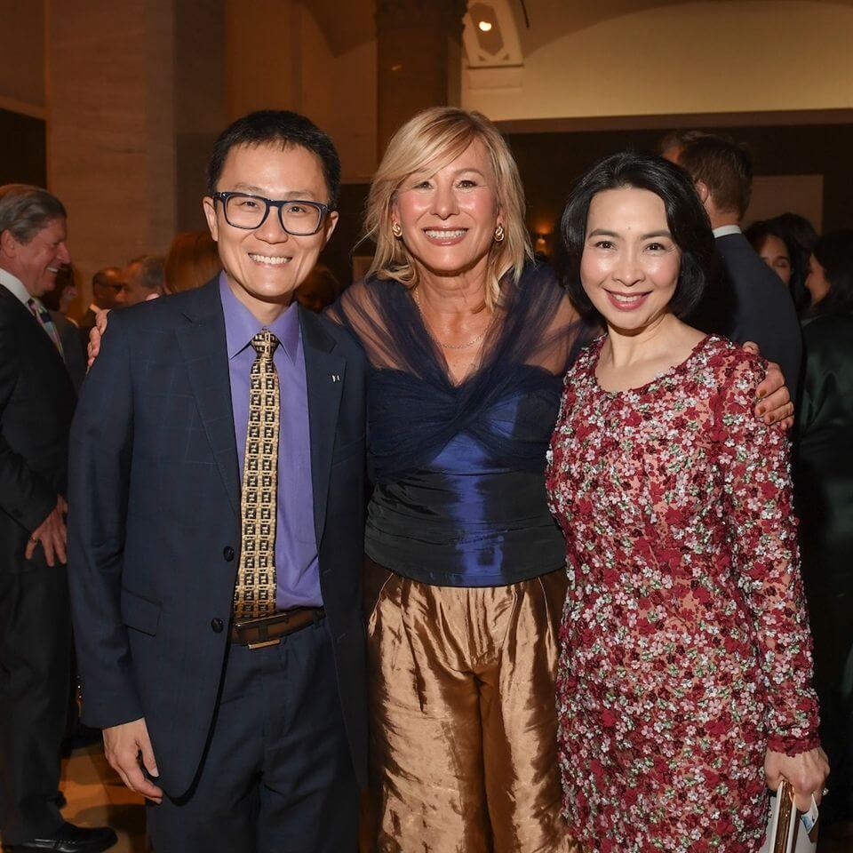 Three people dressed in formal attire stand close together and smile at the camera at a crowded indoor event. The background shows other guests mingling in a warmly lit room.